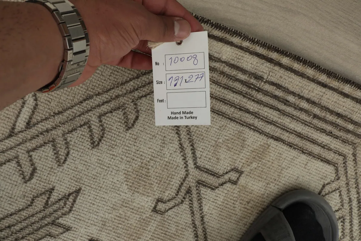 A hand holding a size tag (191x277 cm) over the rug, showing the hand-knotted texture.