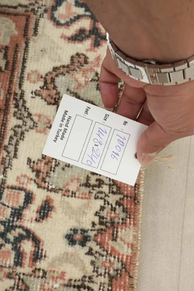 A hand holding a tag with the rug's dimensions (148x240 cm) and 'Hand Made in Turkey' label.