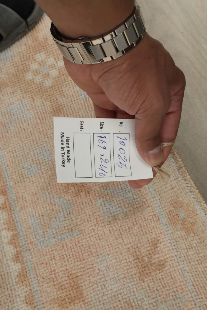 Hand holding a tag showing the rug's dimensions in centimeters and 'Hand Made in Turkey'