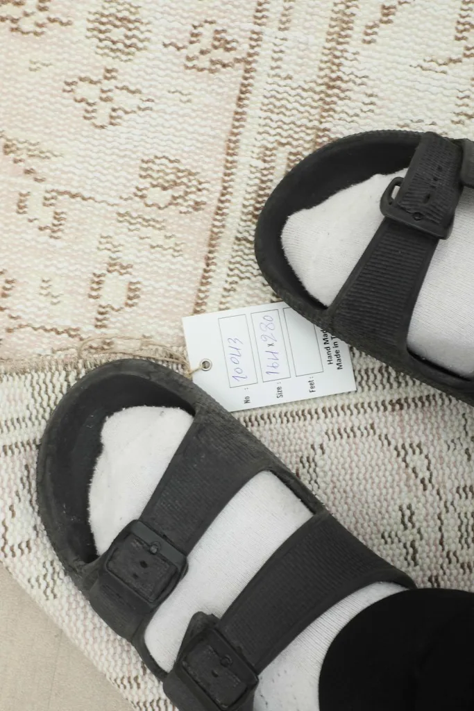 Feet in sandals next to a size tag, providing a sense of scale and texture of the rug's surface.