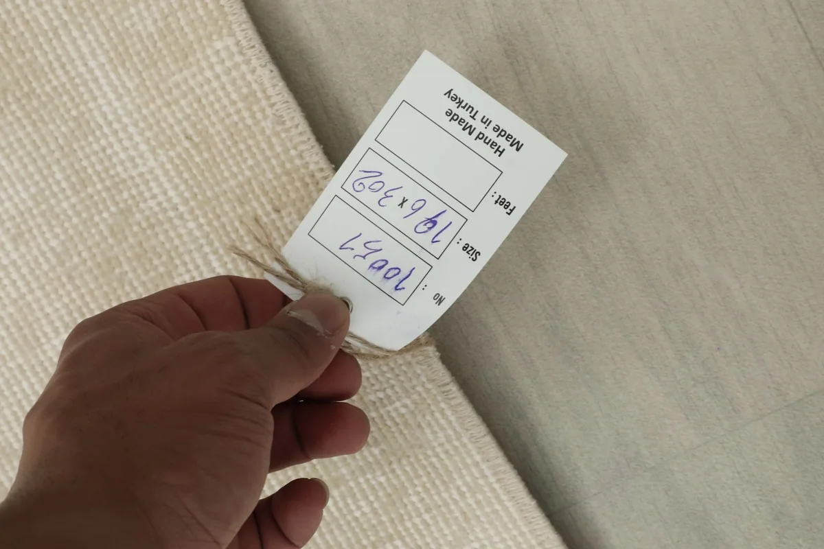 Hand holding the product tag, displaying the rug's dimensions 196x302 cm and origin.