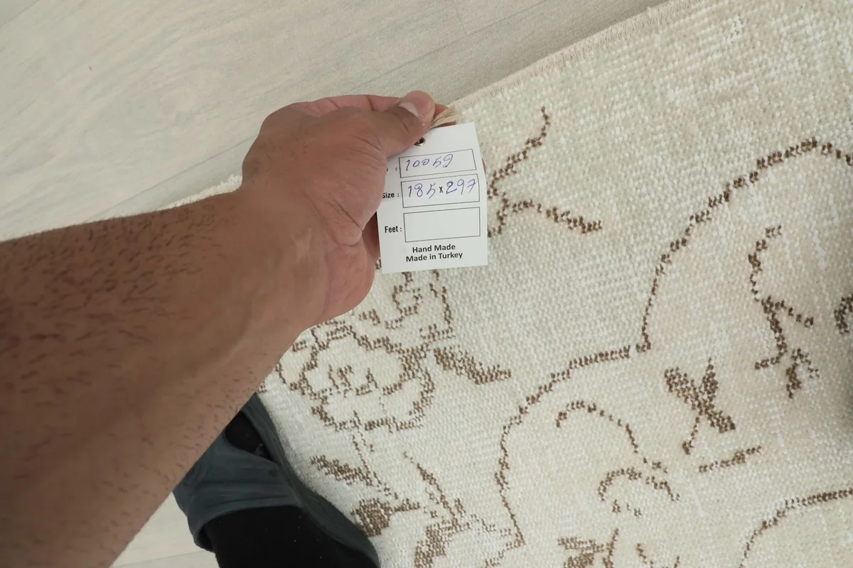 Hand holding a size tag over the rug, showing the 185x297 cm dimensions