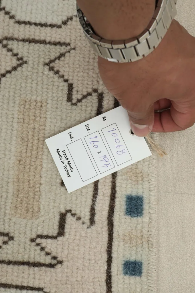Hand holding a tag showing the rug's dimensions in cm and feet, confirming its hand-made origin in Turkey.