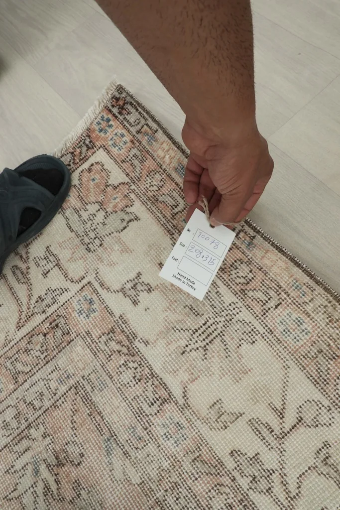 Hand holding a tag near the edge of the rug, showing a close view of the back and knotting.