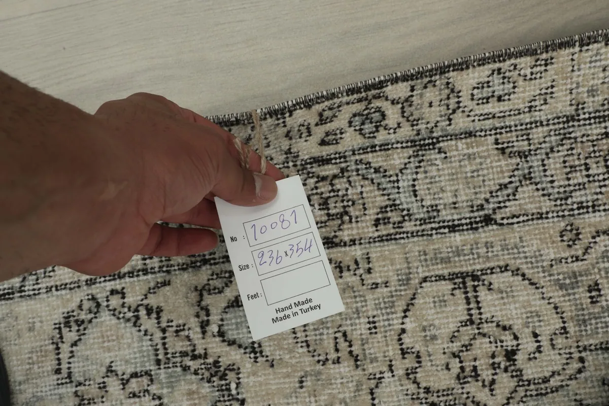Hand holding a tag displaying the rug's unique identifier and dimensions in centimeters.