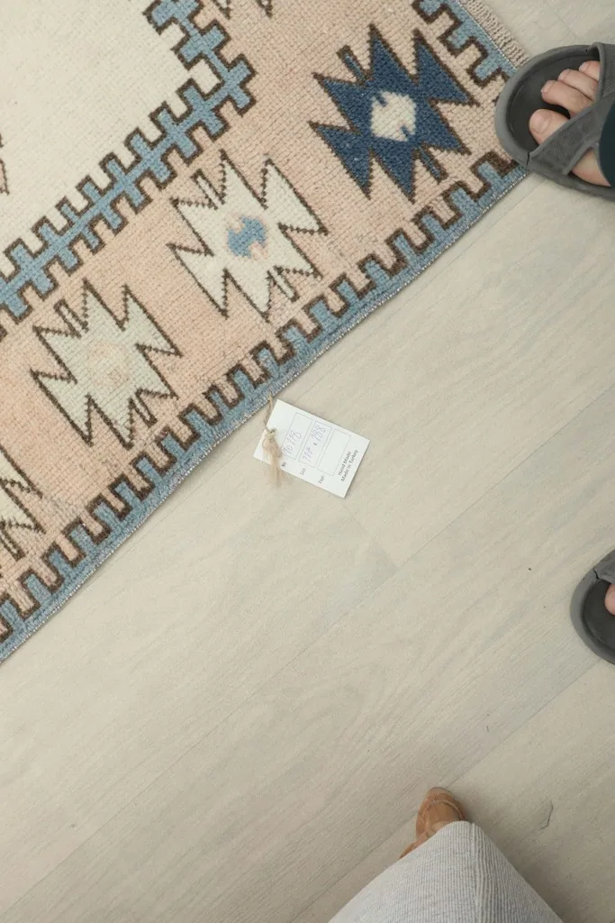A small tag on the floor next to the rug, with feet visible, indicating the rug's size and origin.
