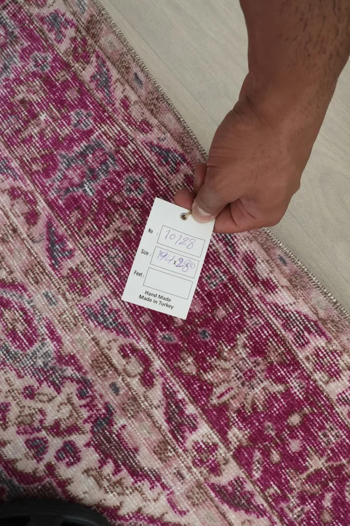 Hand holding a tag on the rug, showing details like 'hand woven' and 'made in turkey'
