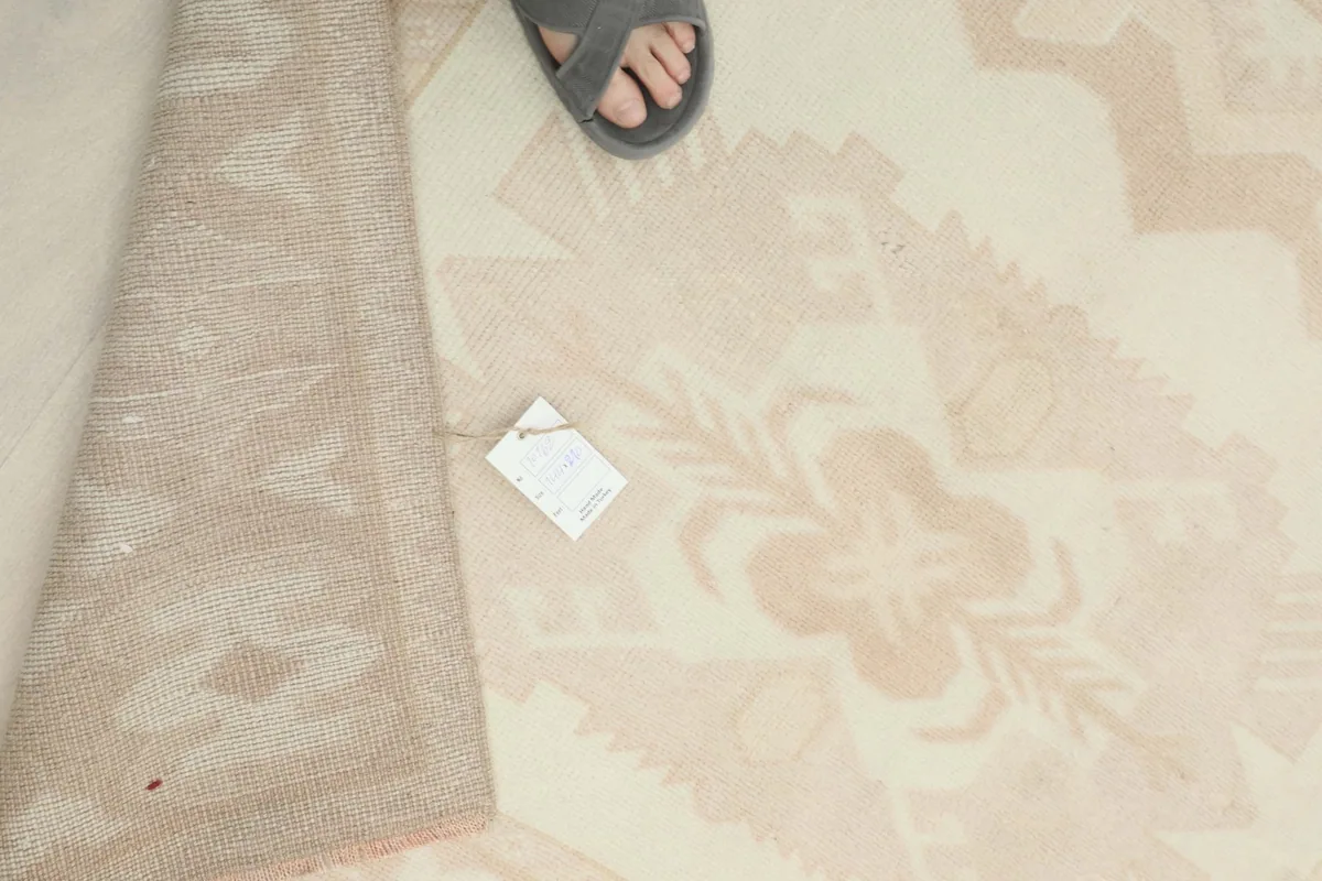 Foot next to the rug with a tag, showing scale and the soft texture of the cream and rose vintage rug.