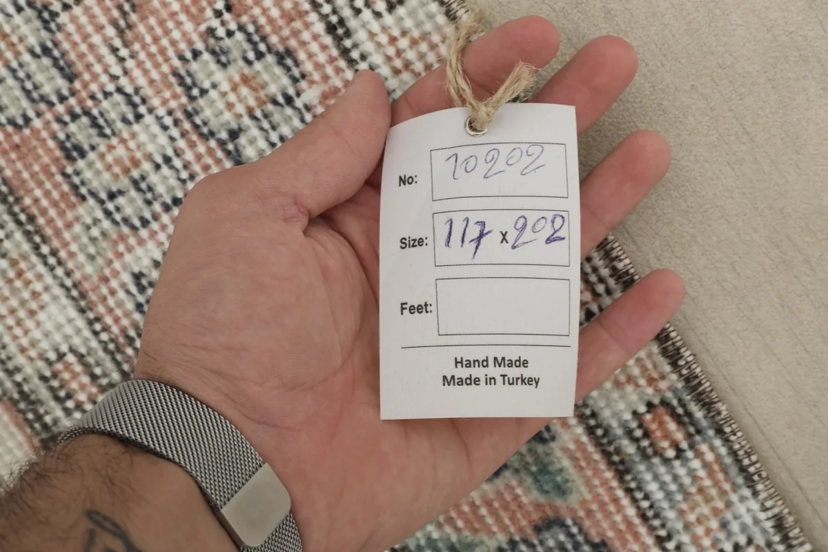 Hand holding a tag showing the rug's number and size in centimeters (117x202), confirming it is handmade in Turkey.