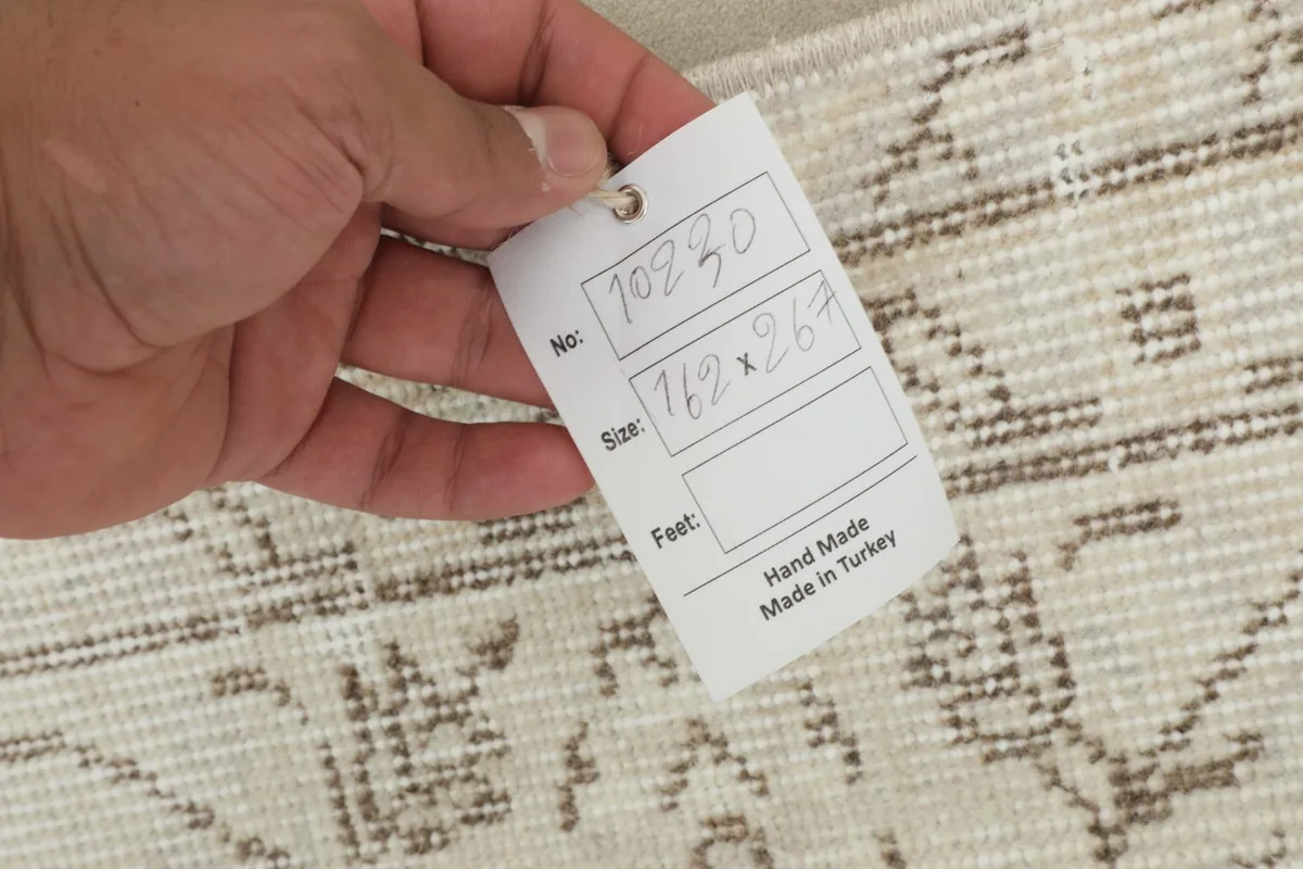 Hand holding a tag showing the rug's size in centimeters and stating 'Hand Made in Turkey'.