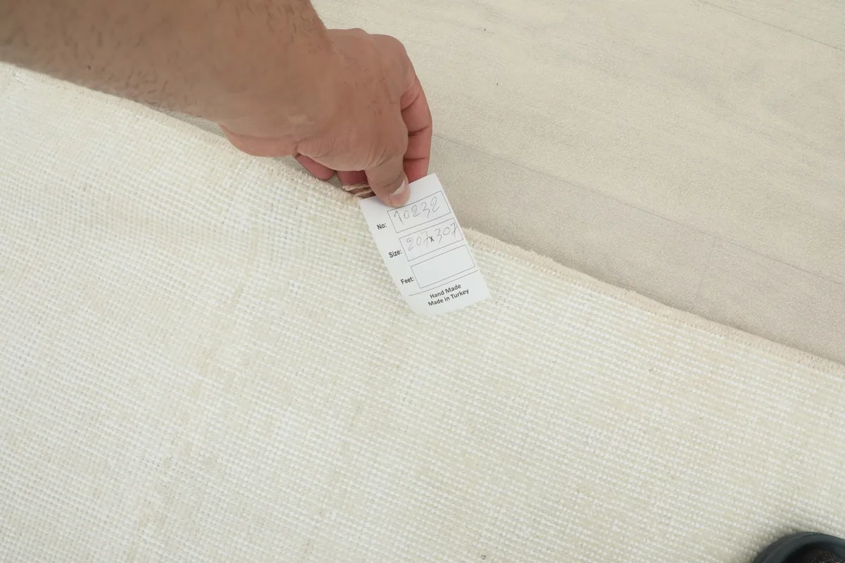 Hand holding a measurement card near the edge of the cream rug, illustrating its pile height and construction.