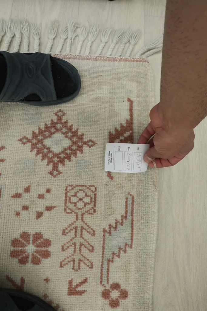 A hand holding a size tag next to the rug, showing scale reference with a shoe