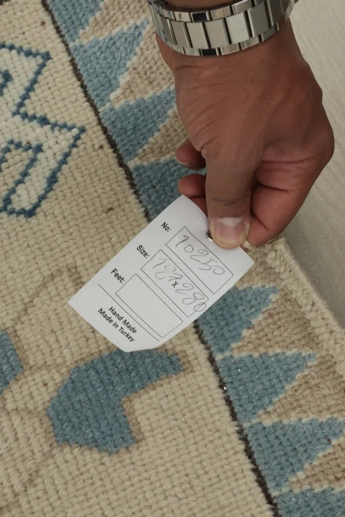 Hand holding a tag showing the rug's size in feet and centimeters, confirming it's handmade