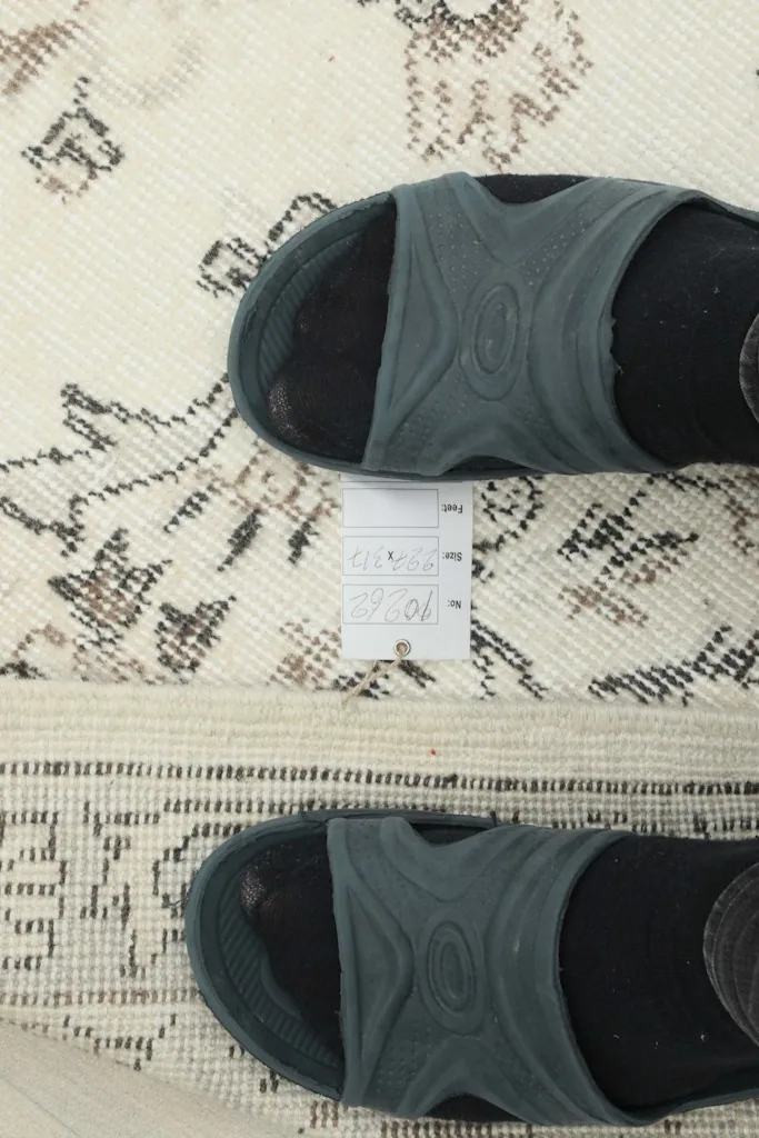 Detail of the rug's low wool pile and a small paper tag with measurements next to a person's feet.
