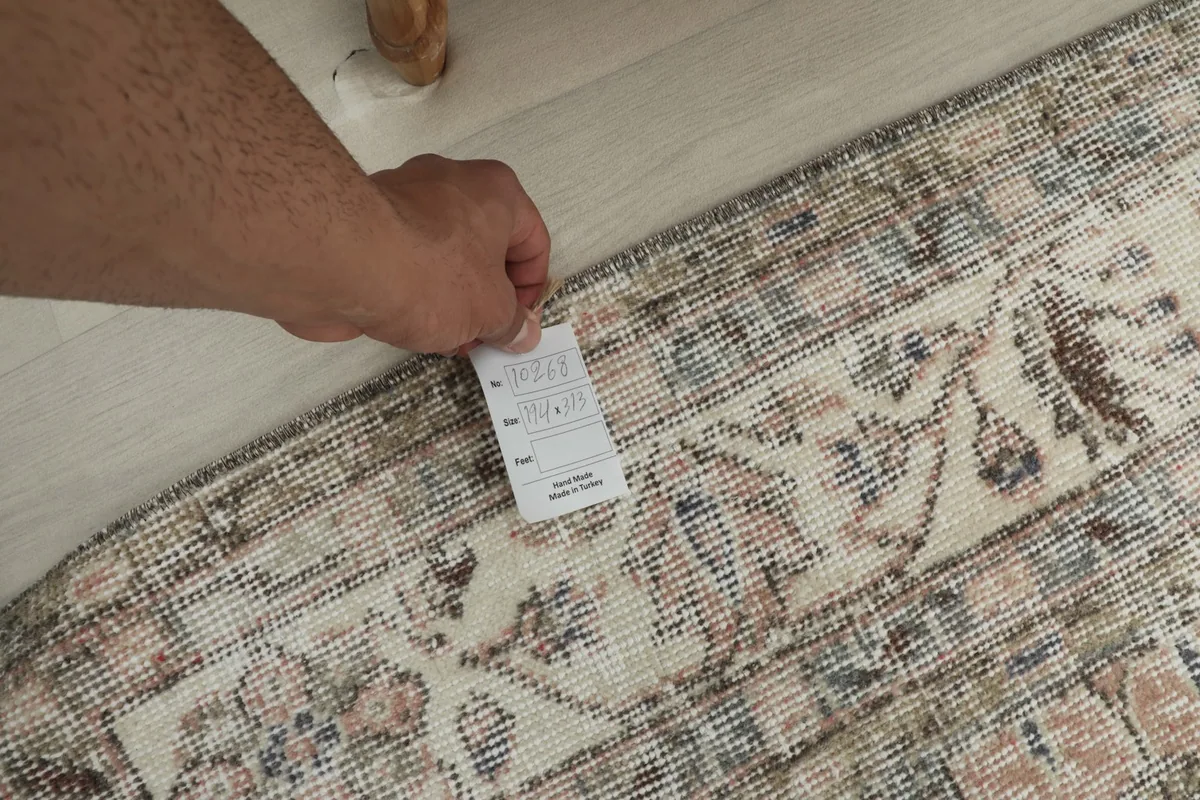 Hand holding a product tag near the rug's edge, showing the backside of the rug and the tag details.