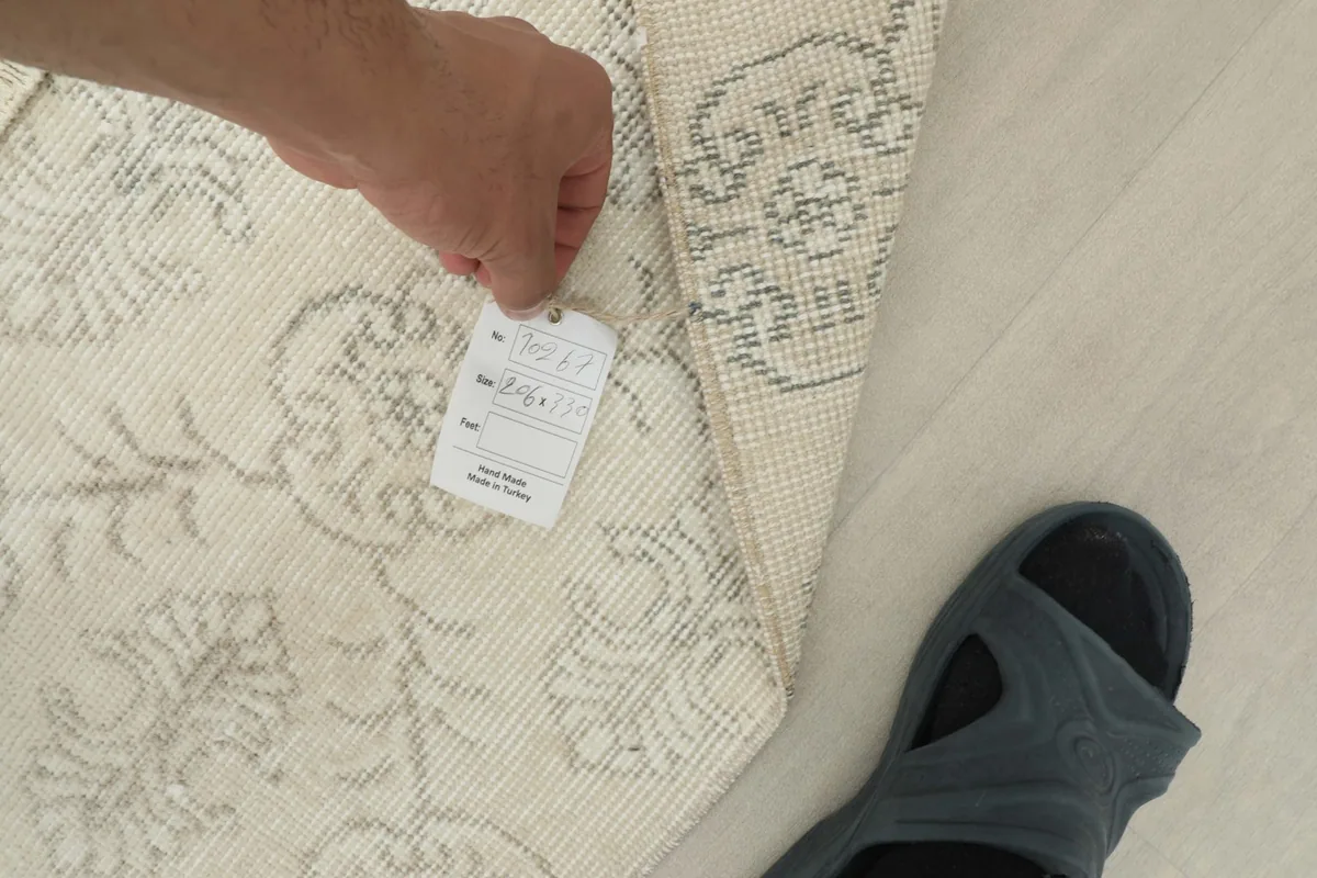 Hand holding a tag near the back of the rug, showing details of its construction and origin.
