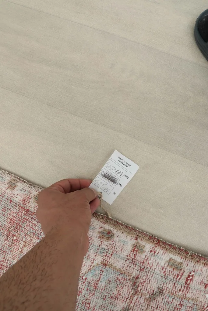 Hand holding a product tag with size and details, next to the edge of the rose area rug.