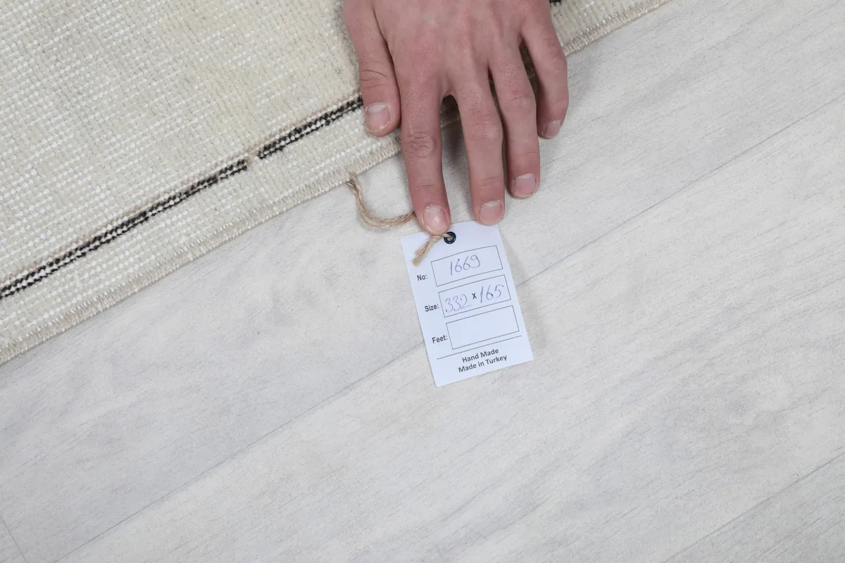 Hand holding a tag on the rug, indicating its hand-knotted origin and size details.