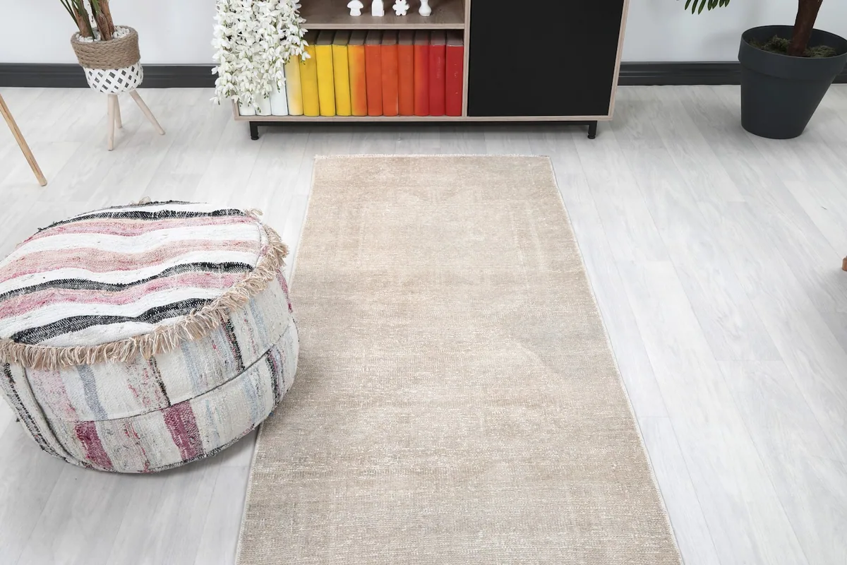 View of the beige runner rug from the end, with an ottoman and shelving unit in the background.