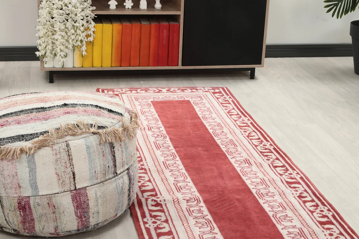 Close-up view of the red runner rug in a room, showing its texture near a pouf and cabinet.