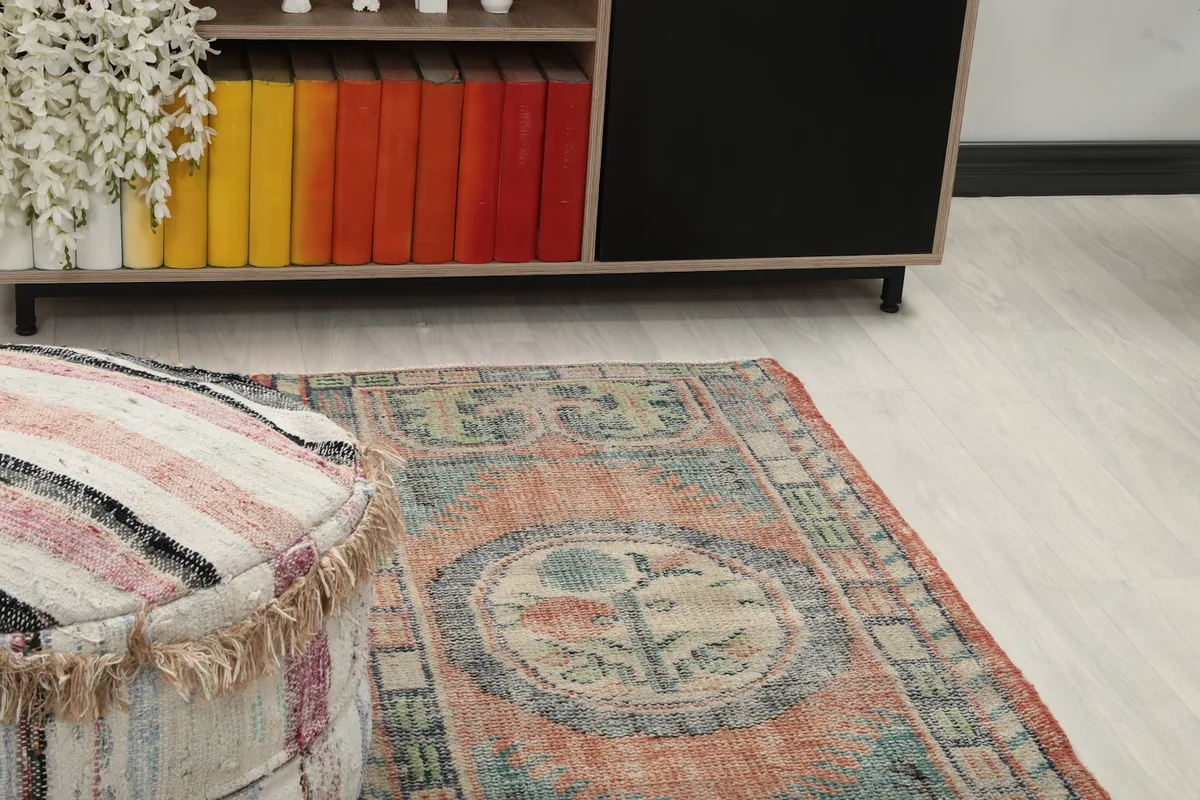 Angled view of the rust and blue rug on a light floor, next to a striped ottoman and a wooden cabinet.