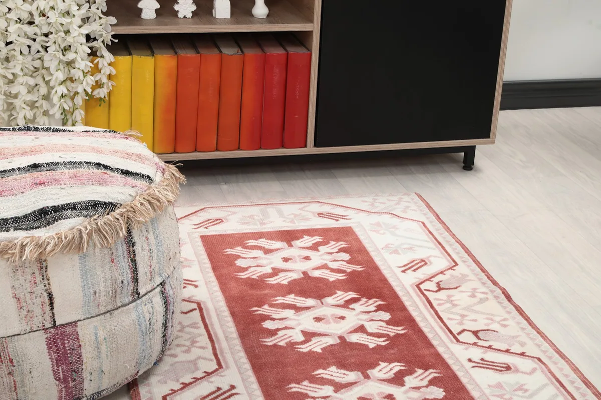 Close-up angle of the rug showing its red field, ivory border, and geometric motifs near an ottoman