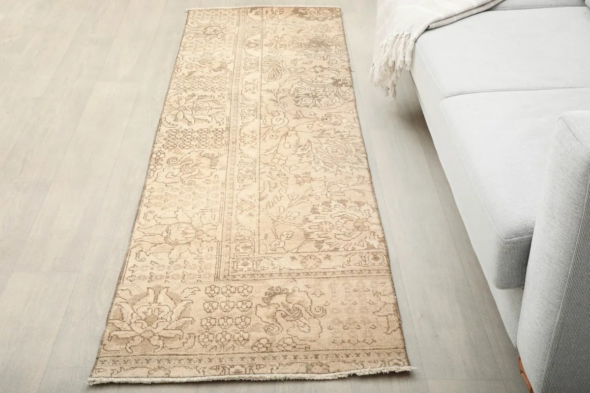 Vintage beige runner rug placed on a light wood floor next to a light gray sofa in a living room setting.