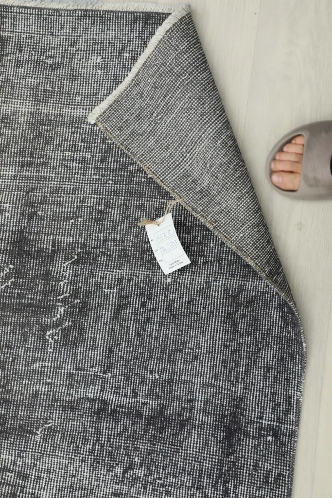 Detail of the rug's back with a label, next to a foot for scale, showing the intricate weaving.