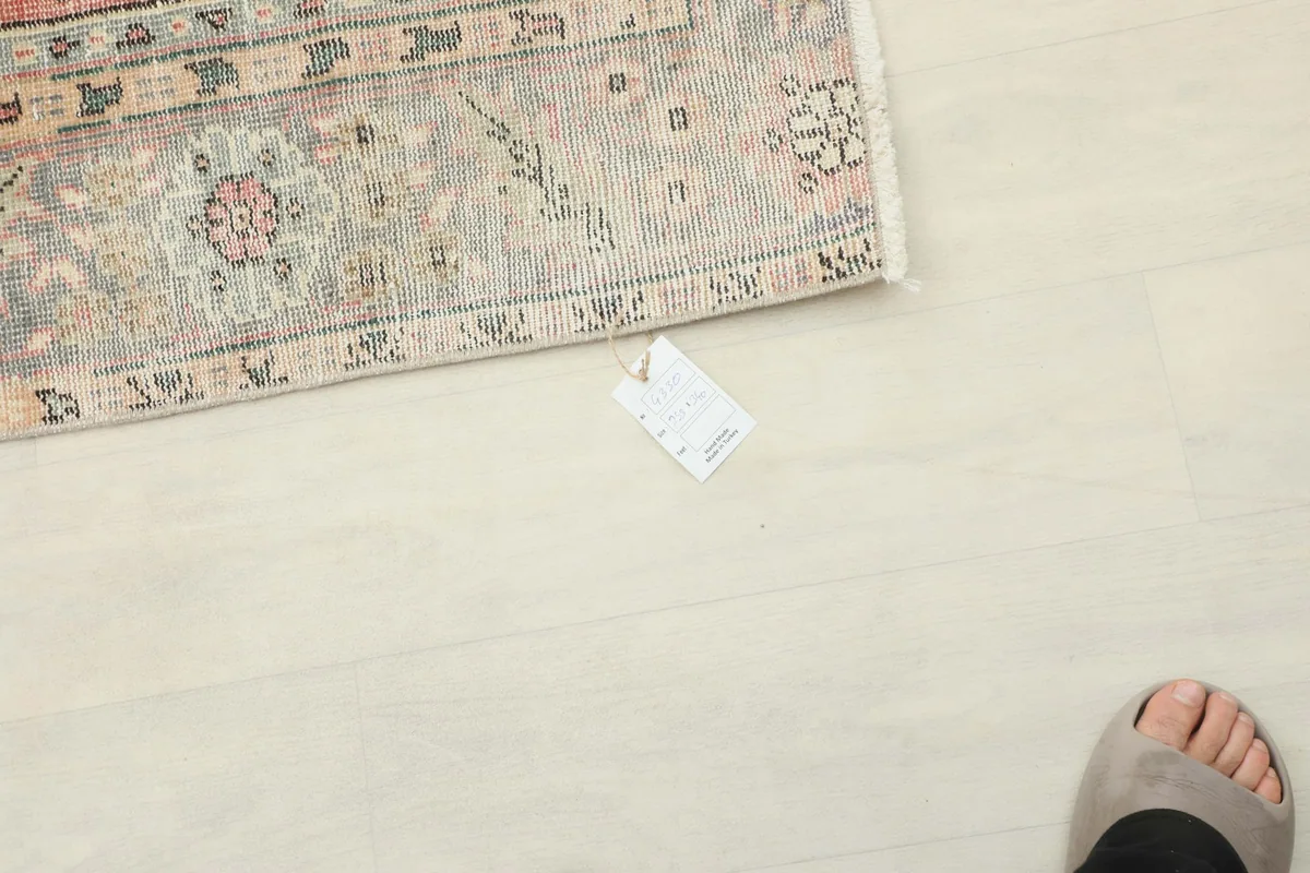 View of the rug's edge and tag on a light floor, with a person's foot nearby for scale