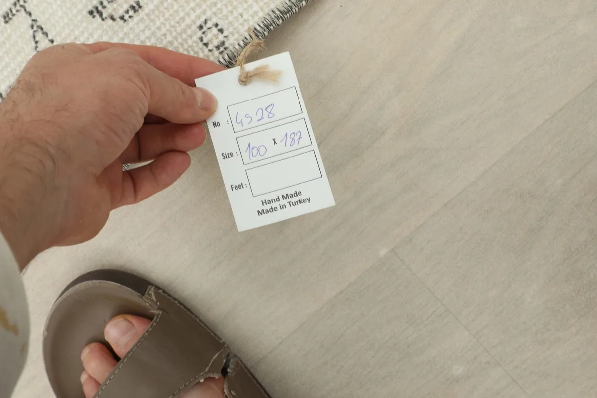 Hand holding a tag with rug details: item number, size in cm, and 'Hand Made in Turkey' text.