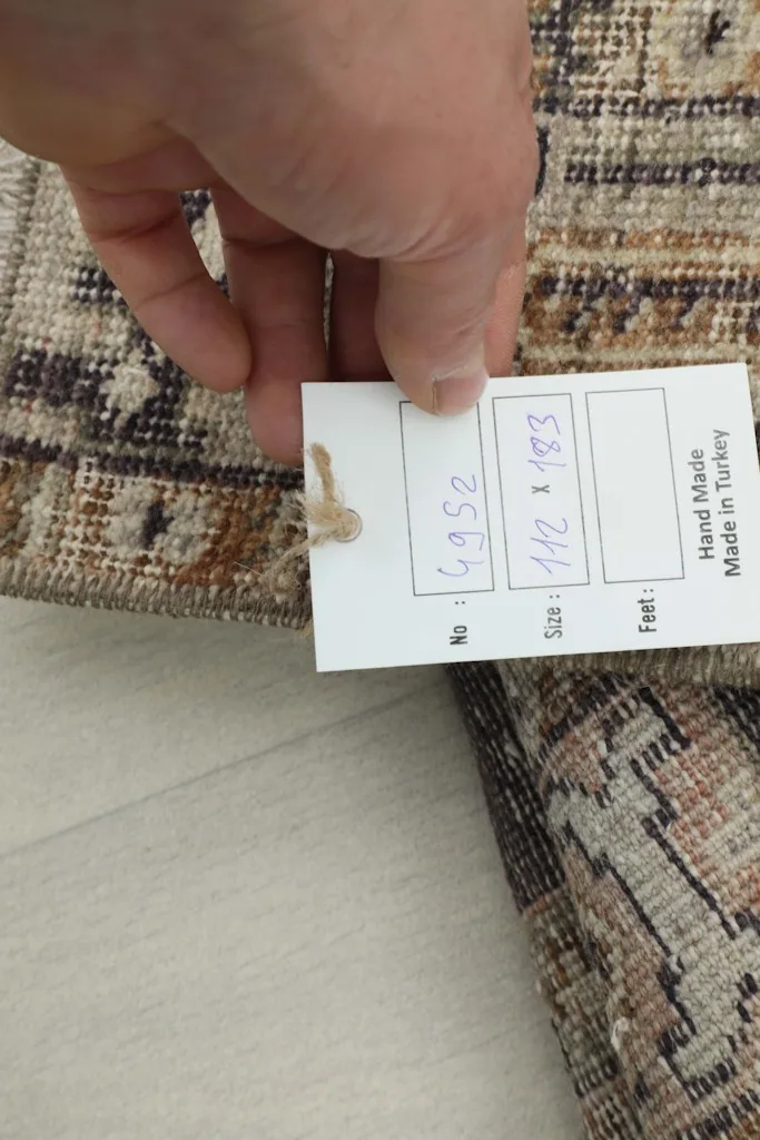 Hand holding a tag showing the rug's size (112 x 183 cm) and confirming it is hand-made in Turkey.