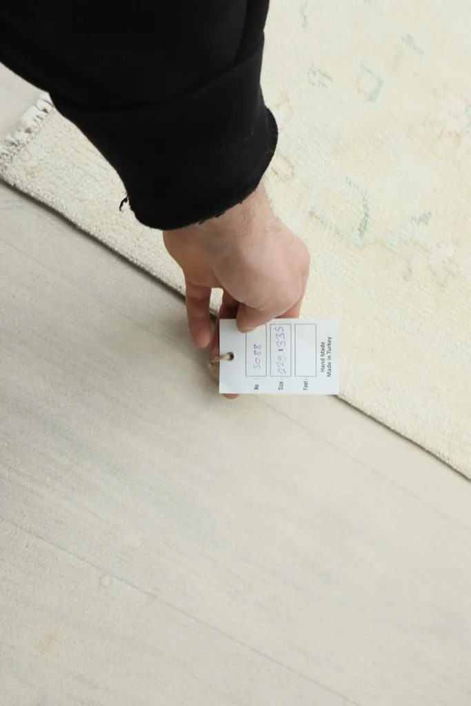 Hand holding a product tag against the rug, showing details like size 7'6" x 11'0"