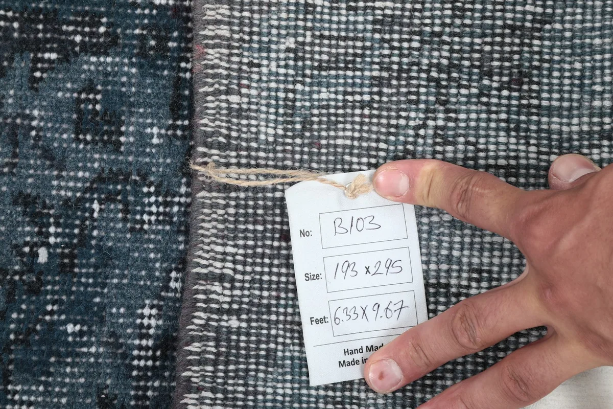 Hand holding a tag showing the rug's dimensions in centimeters and feet, confirming its origin and material.