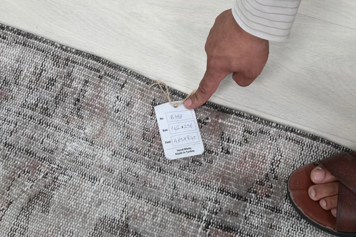 Hand pointing to the product tag on the rug, showing details like size and material