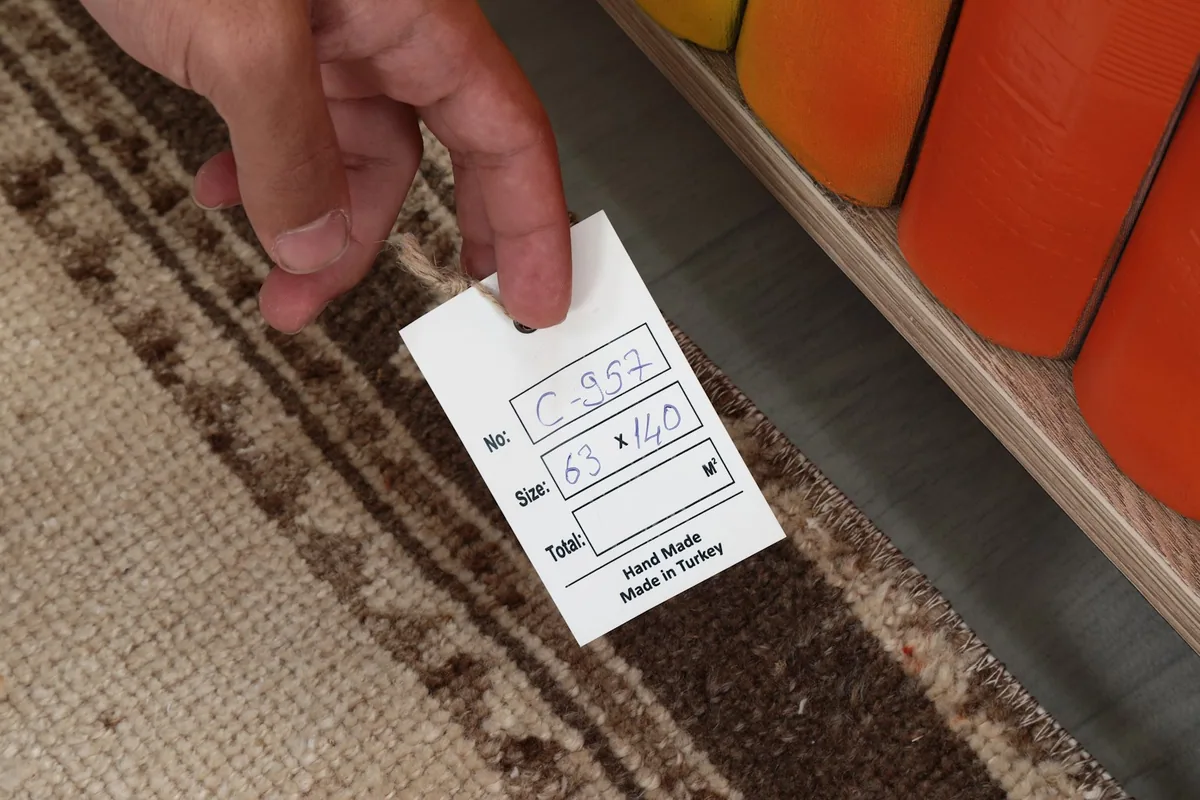 Hand holding a tag displaying the rug's dimensions (63 x 140 cm) and 'Hand Made in Turkey'.