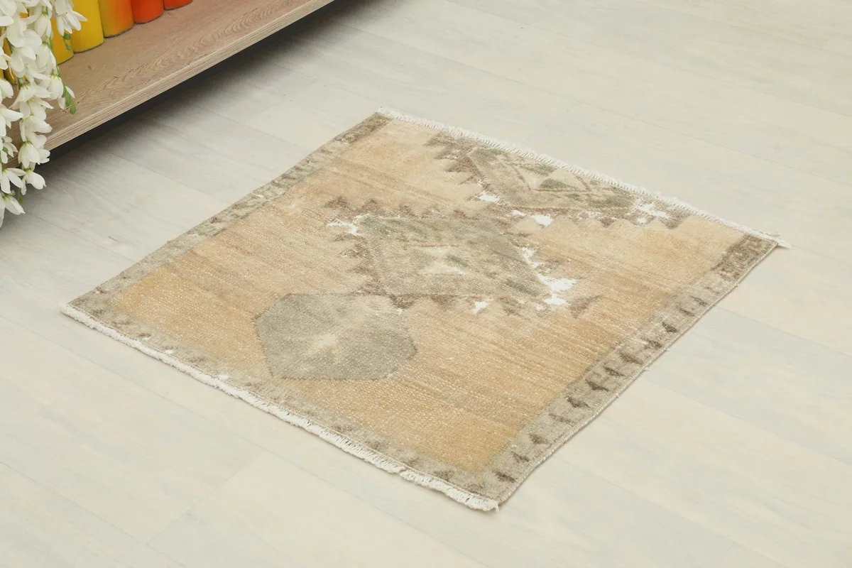 Angled view of the small tan and gray rug, showing its square shape and low pile.