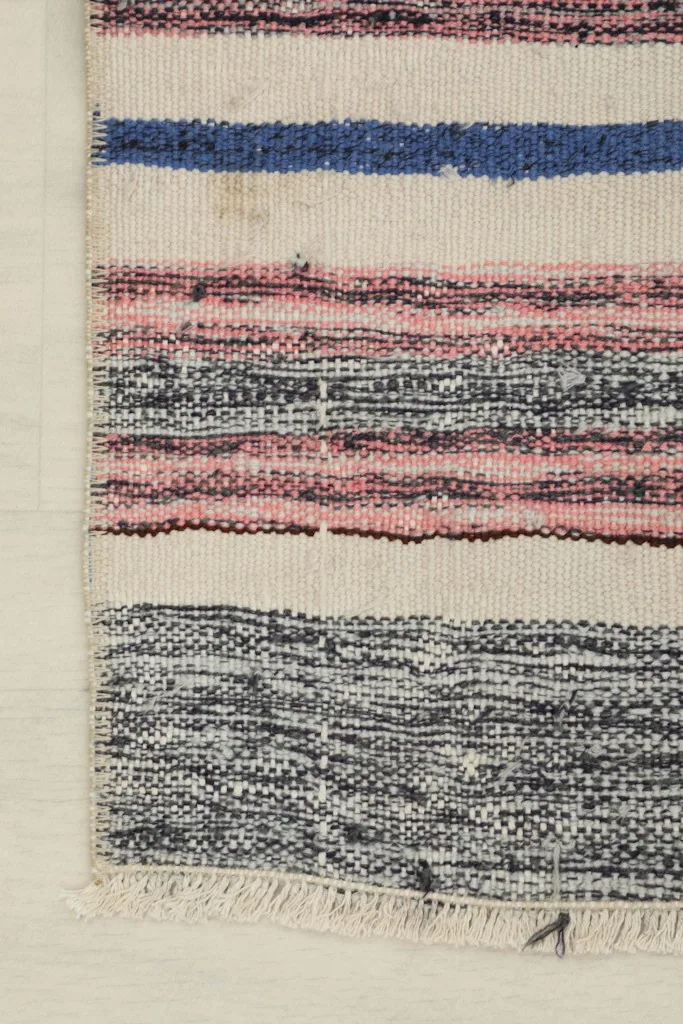 Detail view of the rug's corner, highlighting blue, pink, and charcoal stripes with fringe.
