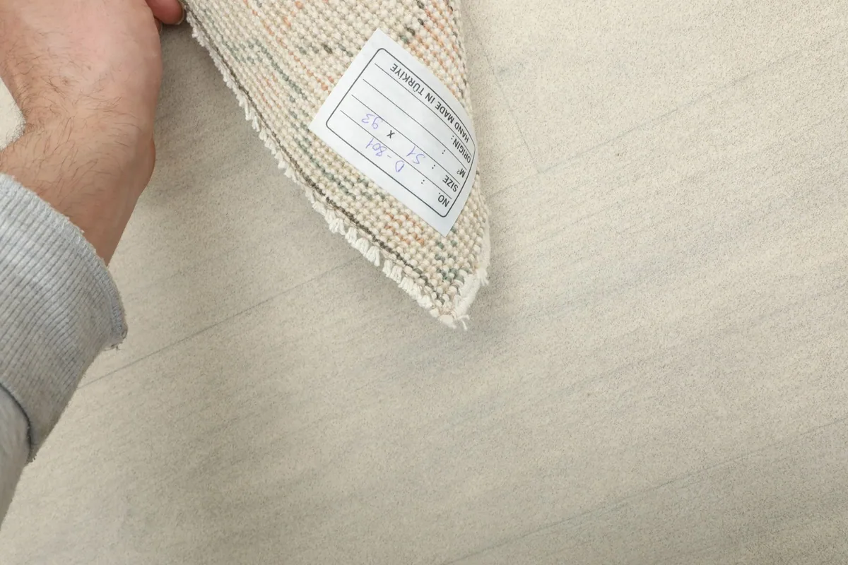 Hand holding a corner of the rug, with a label indicating its origin and dimensions.