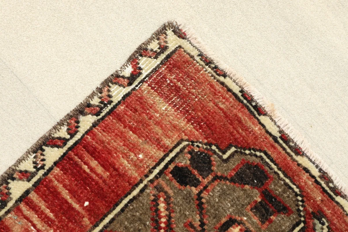 Corner detail of the rug, illustrating the red field, brown geometric patterns, and the narrow ivory and black border.