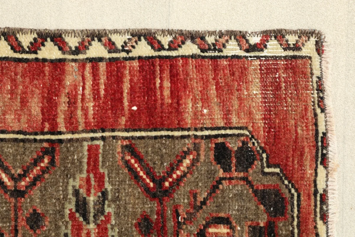 Edge detail of the rug, showing the red main field, brown pattern, and the subtle ivory and black border design.