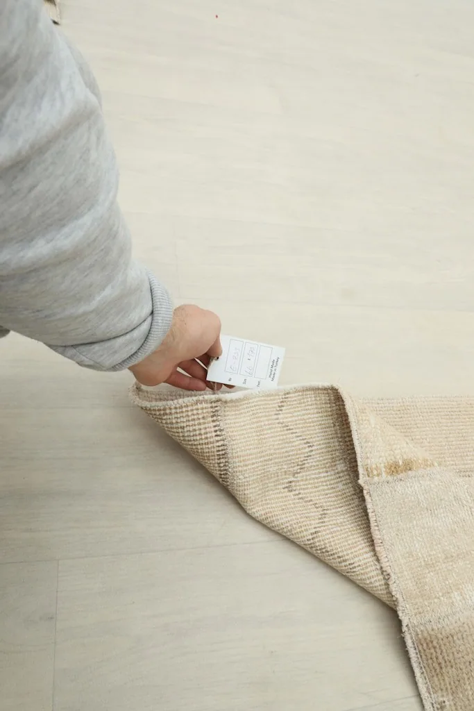 A hand holding a care tag near the folded edge of the rug, showing the reverse side and cotton foundation.
