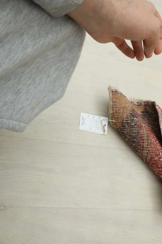 A hand next to the rug with a small tag, indicating the rug's authenticity and origin.