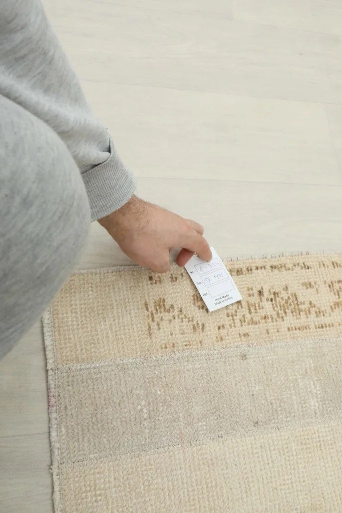 Hand holding a product tag near the rug, showing its texture and construction
