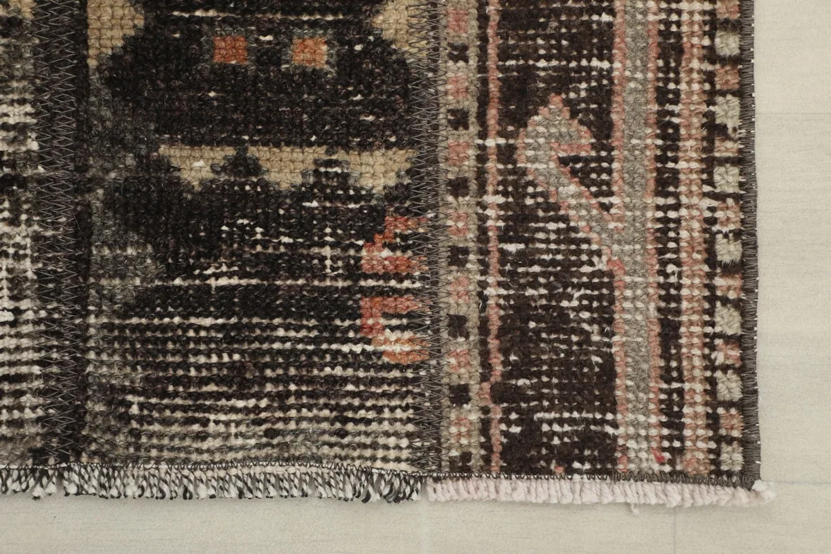 Detail of one end of the rug, showing the faded charcoal pile, beige patterns, and the short, worn fringes.