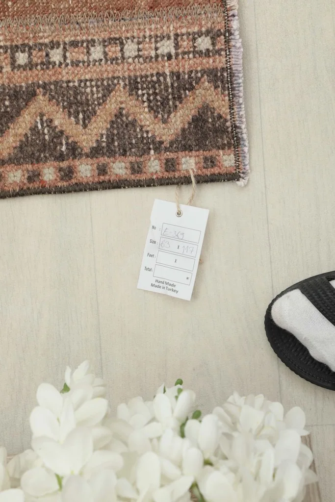 Close-up of the rug's edge with a product tag, near white decorative flowers and a shoe.