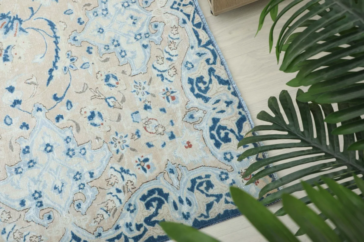 Corner detail of the rug with blue border and beige field, next to green foliage.