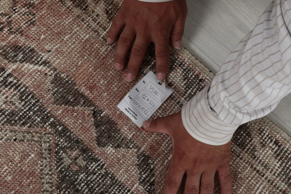 Hands holding a tag on the rug, showing the texture and detail of the wool pile.