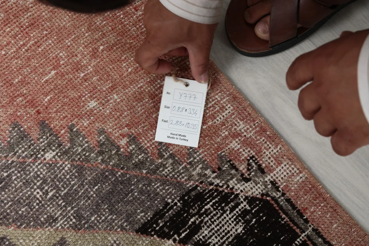 Hand holding a tag, showing the rug's label details and the texture of the wool pile